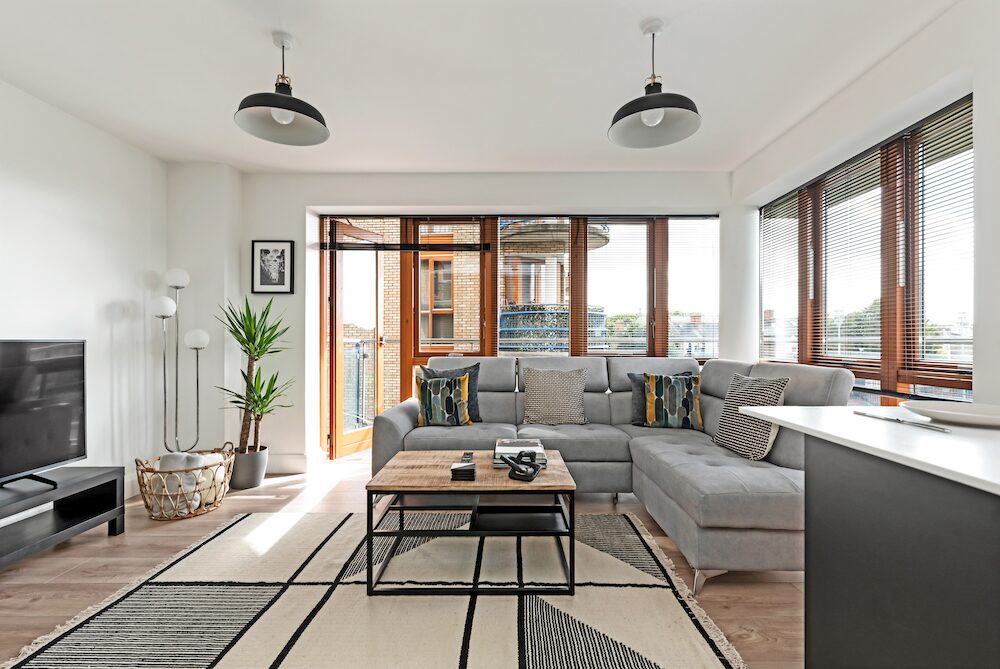 Bright open-plan living room in an EirStay Kilmainham serviced apartment, Dublin 8 — grey sectional sofa, industrial pendant lighting and balcony doors over the city