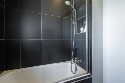 Bath with an overhead shower, glass screen and dark wall tiles at Sandford Gardens
